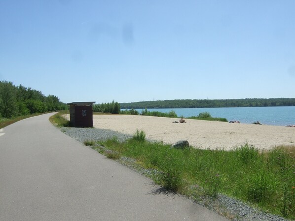 Eine asphaltierte Straße biegt sich nach rechts und führt zu einem Sandstrand, auf dem sich eine kleine Struktur, vermutlich eine Toilette, befindet. Im Hintergrund ist ein Gewässer zu sehen.