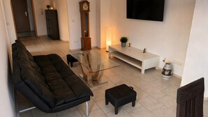 A modern living room with a sofa, ottoman, glass coffee table, TV, and a clock. - Apartment 'Control Suites Og Rechts' with Private Terrace and Wi-Fi (Urbar)