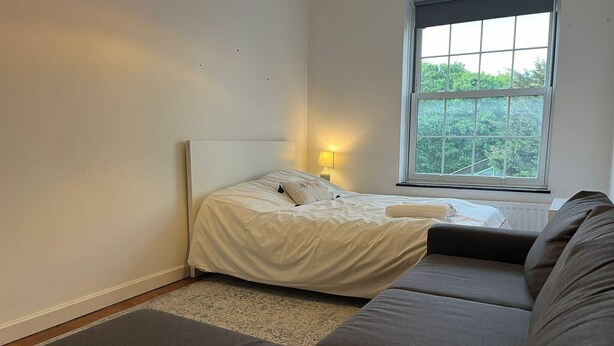 Un dormitorio con cama, lámpara de noche, ventana y sofá.