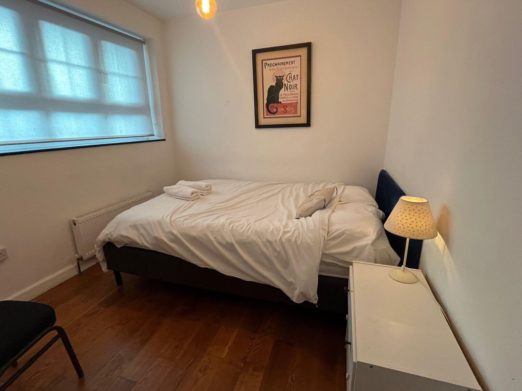 A bedroom with a bed, a desk with a lamp, a radiator, and a window with blinds.