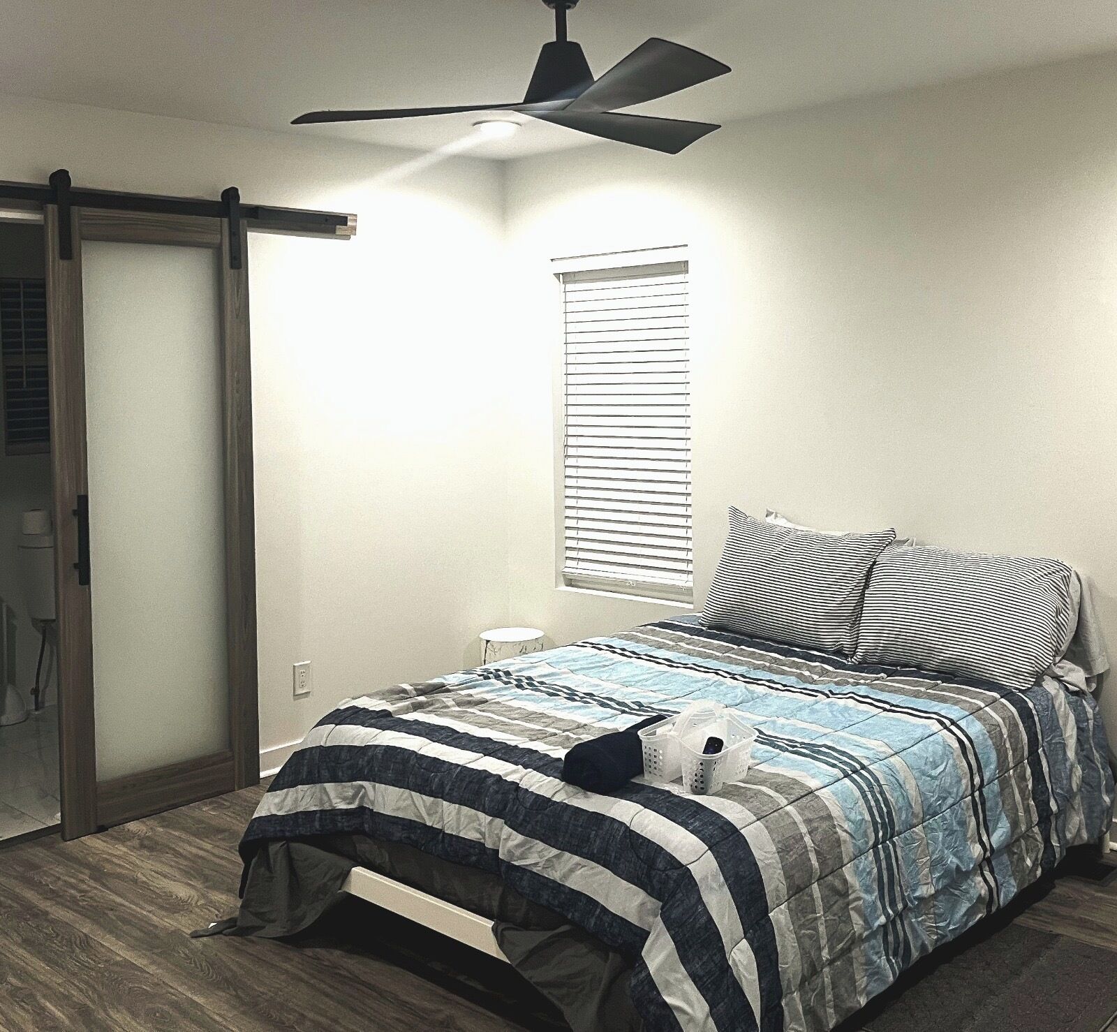 A bedroom with a bed, a ceiling fan, a window with blinds, and a door.