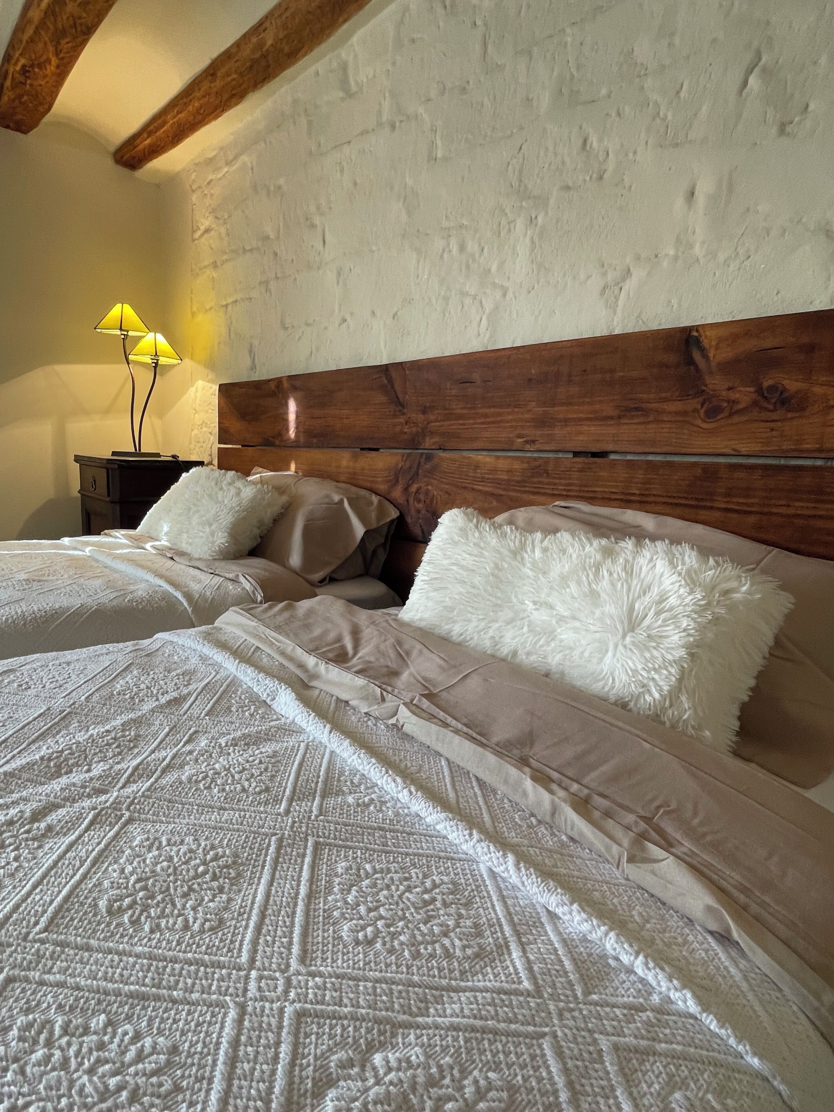 Un dormitorio con cabecera de madera, dos camas con ropa de cama blanca y beige, una mesita de noche con lámpara y una pared con textura.