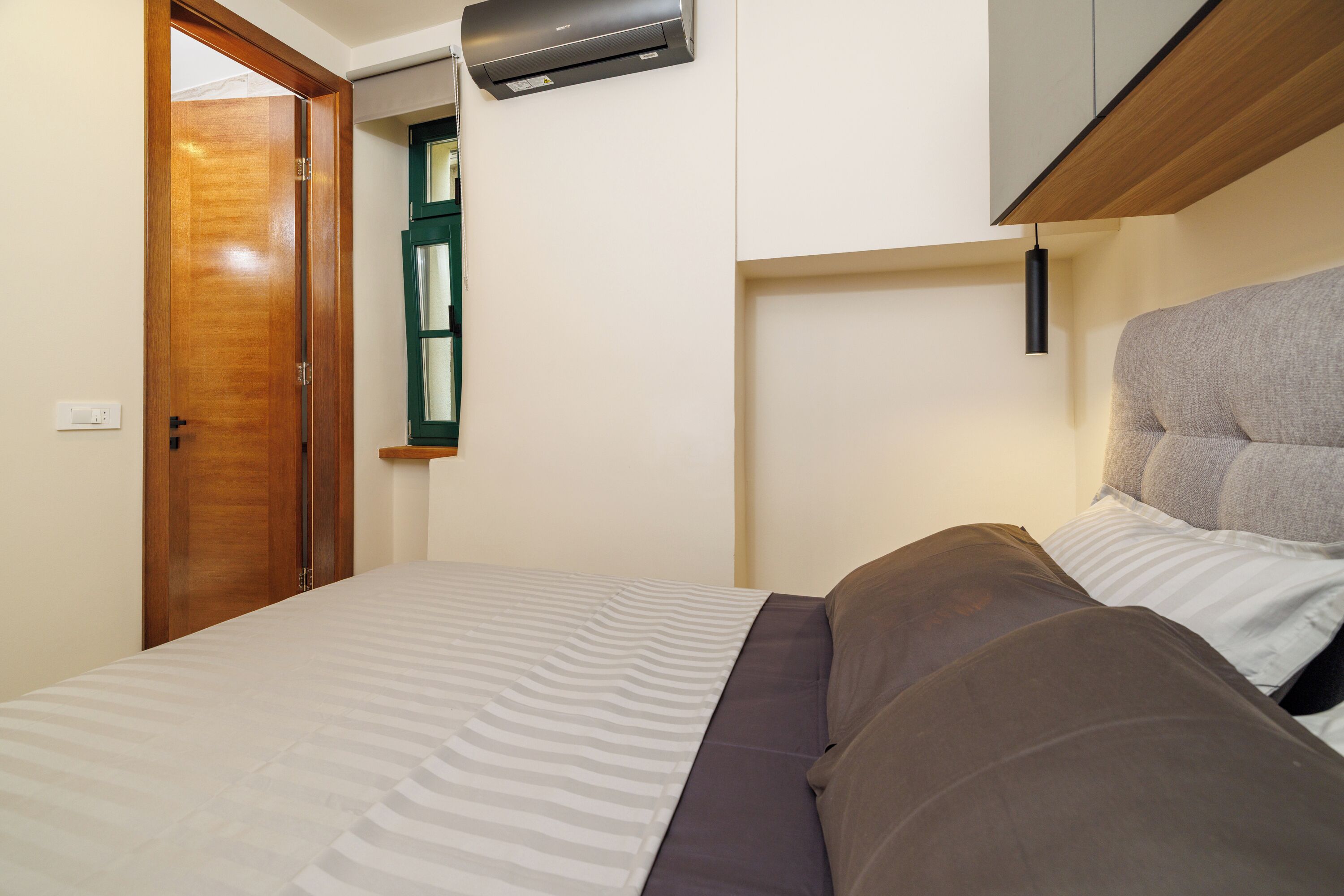Een slaapkamer met een bed, een houten deur, een raam en een luchtverkoeler aan de muur.