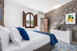 Ein Schlafzimmer mit einem Bett, weißen und blauen Kissen, einem Schminktisch und einer Wand aus Stein, sowie einem Fenster.