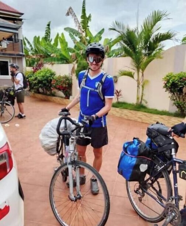 A person standing next to a bicycle with a backpack, in a paved outdoor area with greenery and a building in the background.