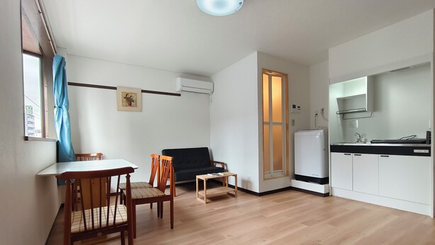 Ein kompakter Wohnbereich mit Esstisch, zwei Stühlen, einer Couch, einem kleinen Tisch, einer Küchenzeile inklusive Kühlschrank und einem Fenster mit Vorhang.