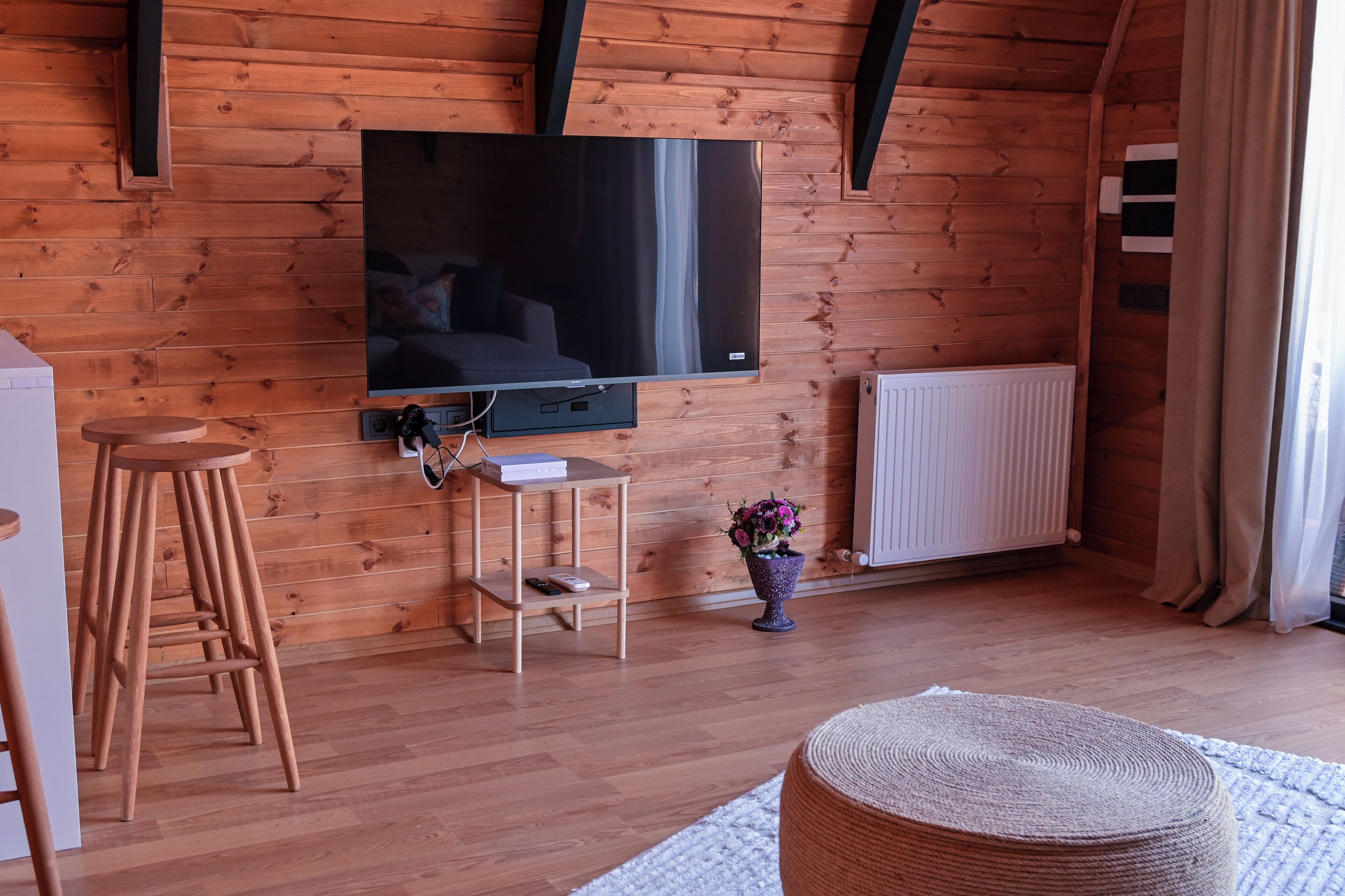 A wooden-paneled room with a flat-screen TV, wooden stools, a small table, a radiator, and a window with curtains.