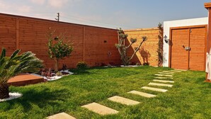 Un patio trasero con valla de madera, un camino pavimentado y un pequeño jardín con plantas y una palmera.