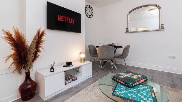 A living room with a TV, a clock, a mirror, a dining table with chairs, and a Scrabble board. - Cosy Home 7 Minutes From Newcastle City Centre (Gateshead)