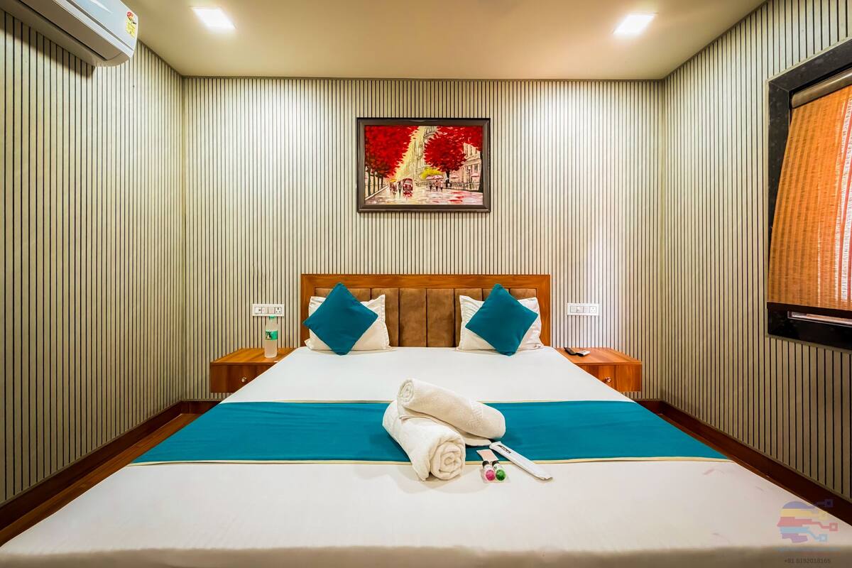 A hotel room with a large bed, a headboard, a painting on the wall, and an air conditioning unit.