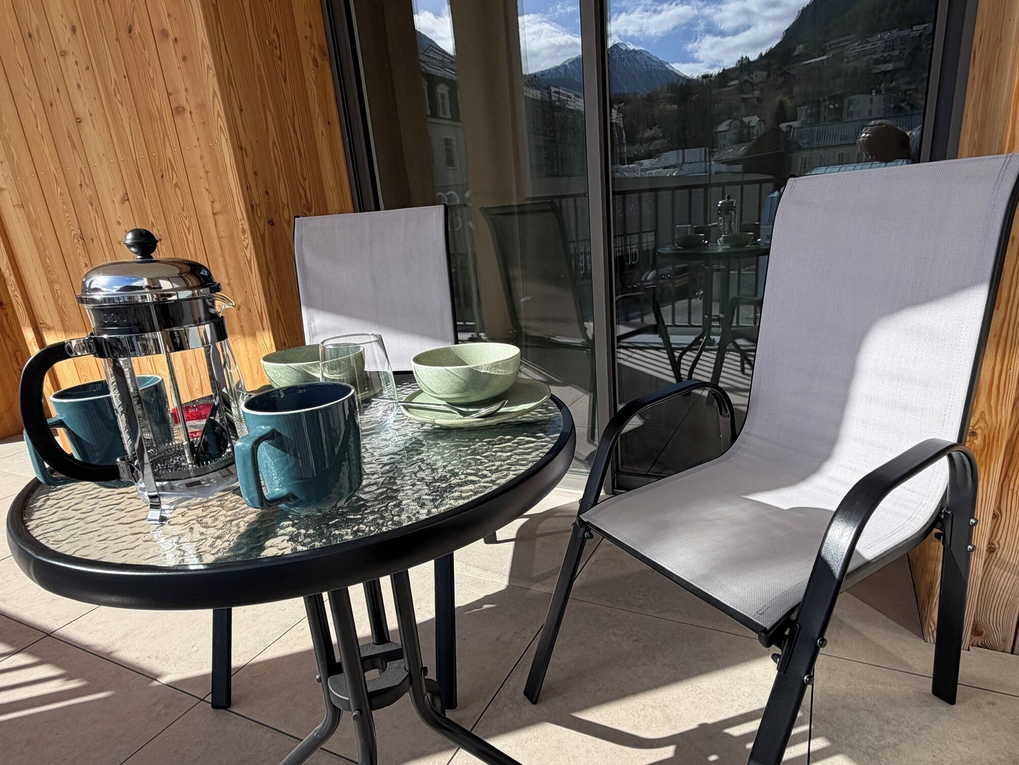 A round table with a glass top, two chairs, a coffee maker, and a view of mountains through large windows.