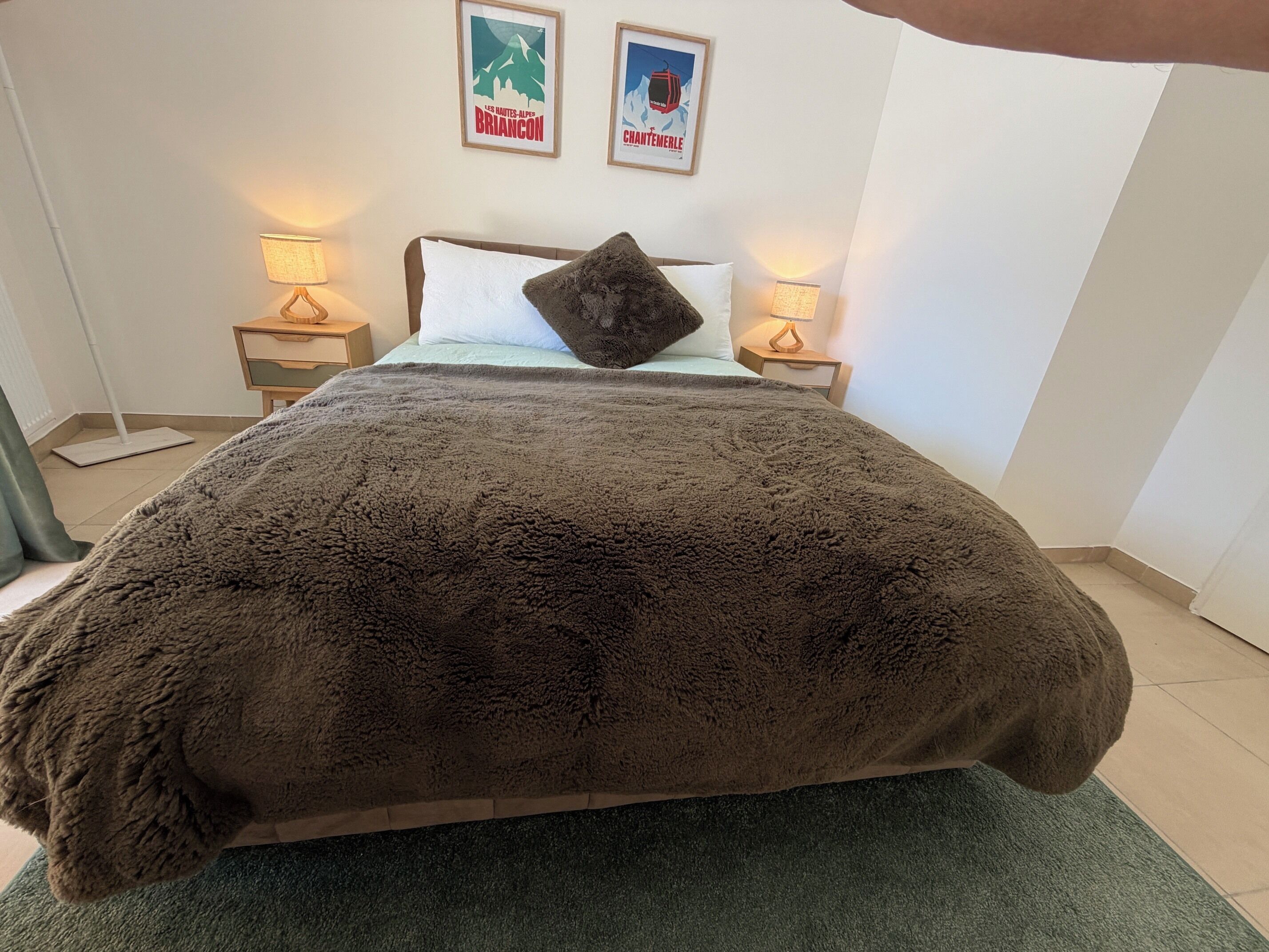Een bed met een bruine deken, twee bedlampen en twee ingelijste foto’s aan de muur.