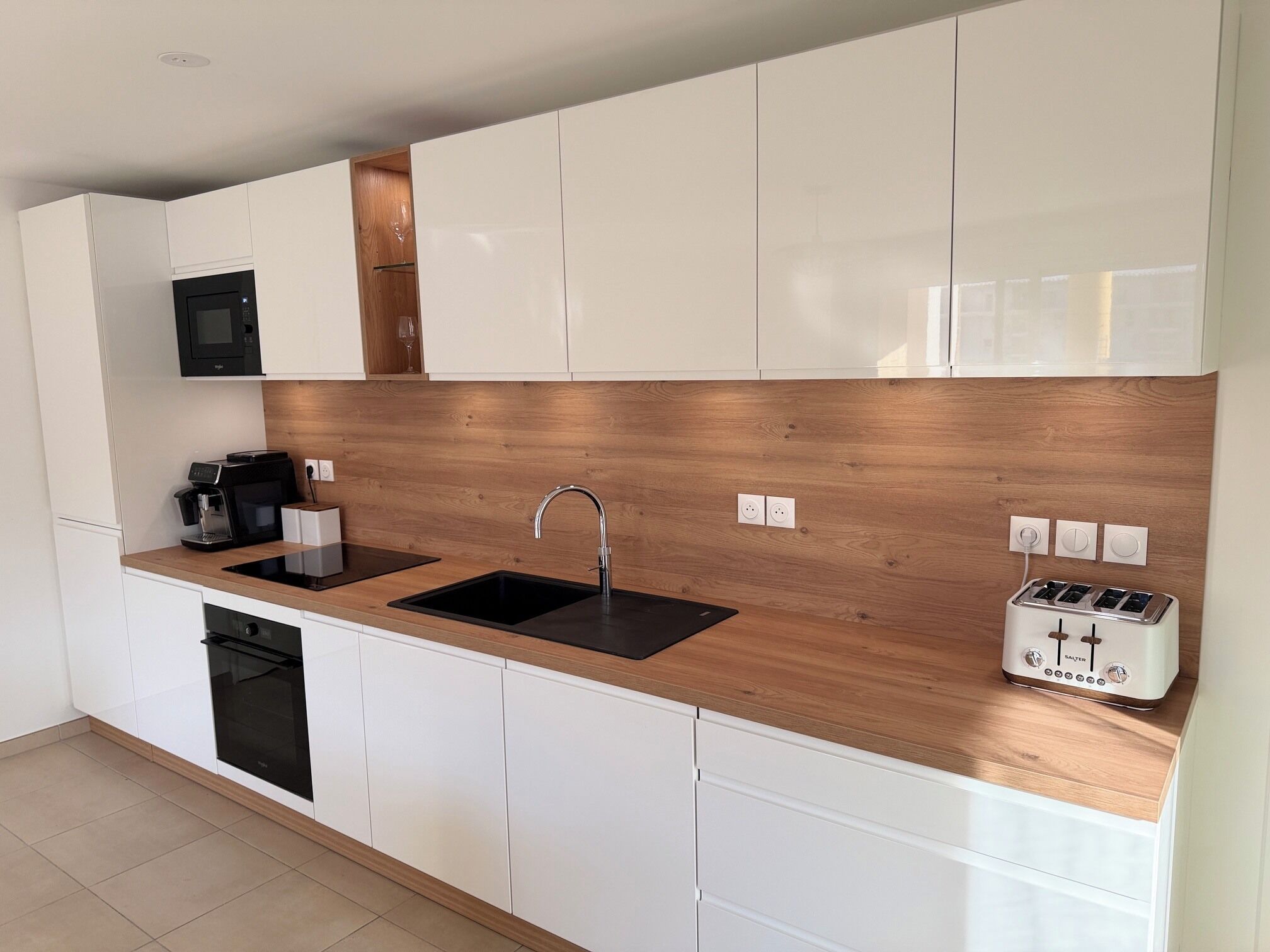 Eine moderne Küche mit weißen Schränken, einer Holzrückwand, einem schwarzen Spülbecken, einer Kaffeemaschine, einem Toaster und einem eingebauten Backofen.