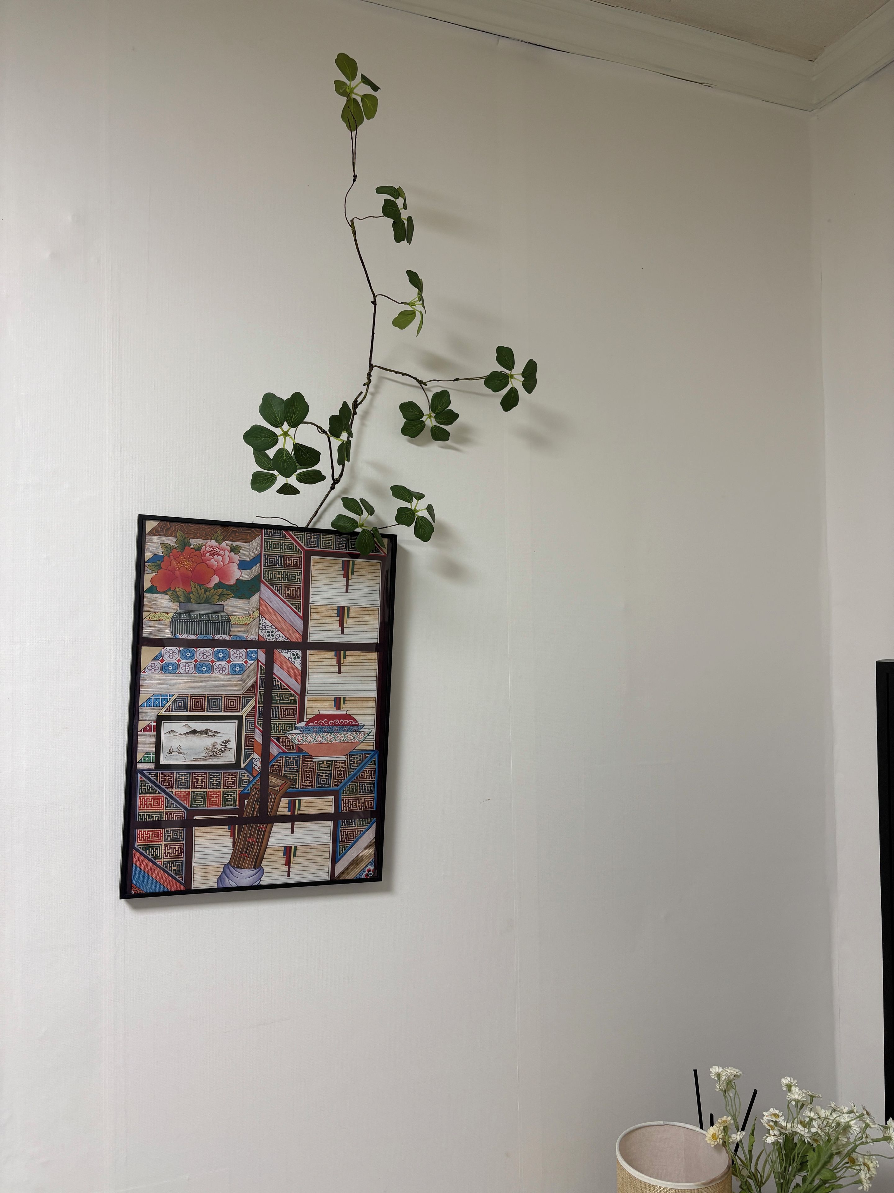 Une œuvre d’art murale dans laquelle une plante pousse, un tableau encadré aux motifs géométriques et une petite plante en pot ornée de fleurs blanches.