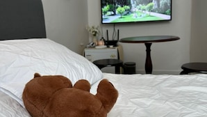 Uma cama com um edredom branco, um urso de pelúcia marrom, uma televisão exibindo uma paisagem e uma pequena mesa com um vaso e um relógio.