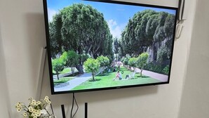 Uma televisão presa na parede, um pequeno eletrodoméstico branco e alguns objetos sobre uma mesa.