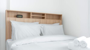 A hotel room with a wooden headboard, white bedding, and a small shelf with a clock. - Free Housekeeping Visits During Your Stay - Stayshort - Peaceful Studio in Jebel Ali That Sleeps 2 (Dubai)
