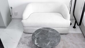 A white sofa, a round marble table, and a patterned rug in a room with white walls and a tiled floor. - Free Housekeeping Visits During Your Stay - Stayshort - Peaceful Studio in Jebel Ali That Sleeps 2 (Dubai)