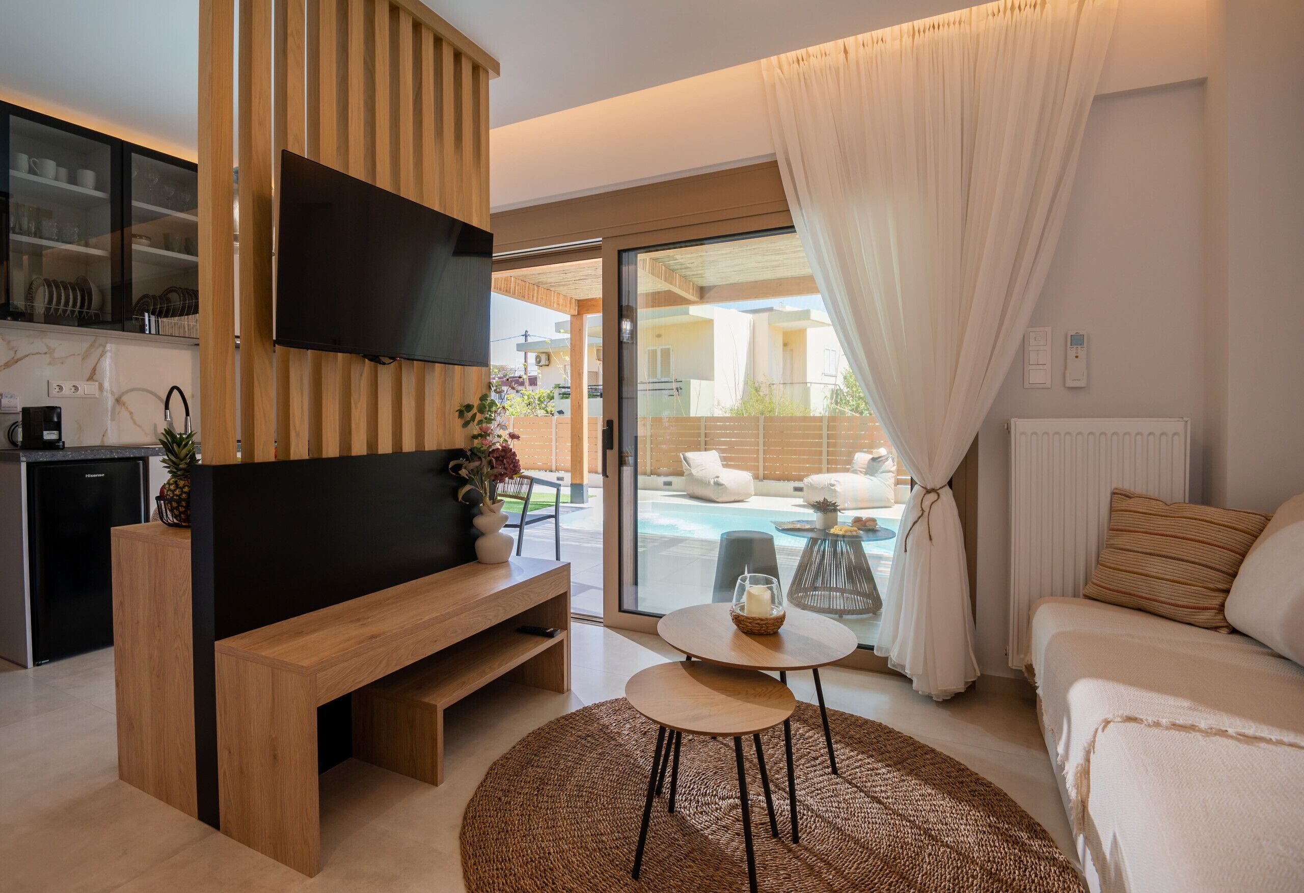A modern living room with a flat-screen TV, wooden furniture, a round table with a candle, and a view of an outdoor swimming pool.