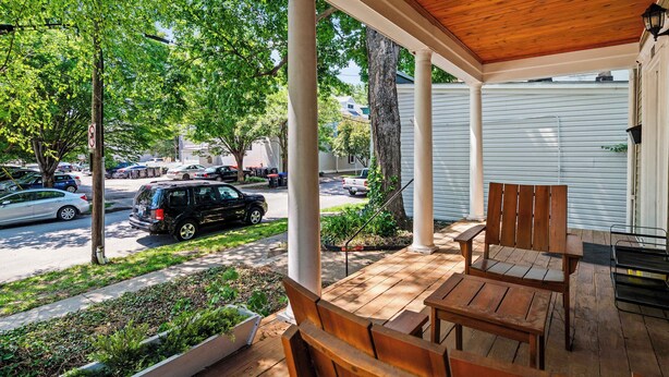 A covered porch with wooden furniture, a view of a street with parked cars, and a tree-lined street.