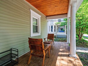 Une véranda en bois avec un banc et des chaises, une colonne blanche et une vue sur une rue bordée de maisons.