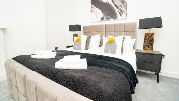 A neatly made bed with a black and beige duvet, two bedside tables with lamps, a large abstract painting on the wall, and a grey carpet. - The Residence by Guestz (Cheltenham)