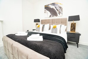 A neatly made bed with a black and beige duvet, two bedside tables with lamps, a large abstract painting on the wall, and a grey carpet. - The Residence by Guestz (Cheltenham)