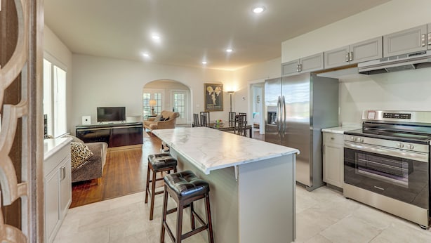 Una cocina moderna con una isla central, taburetes de bar, refrigerador, horno y un área de comedor con sofá y mesa.