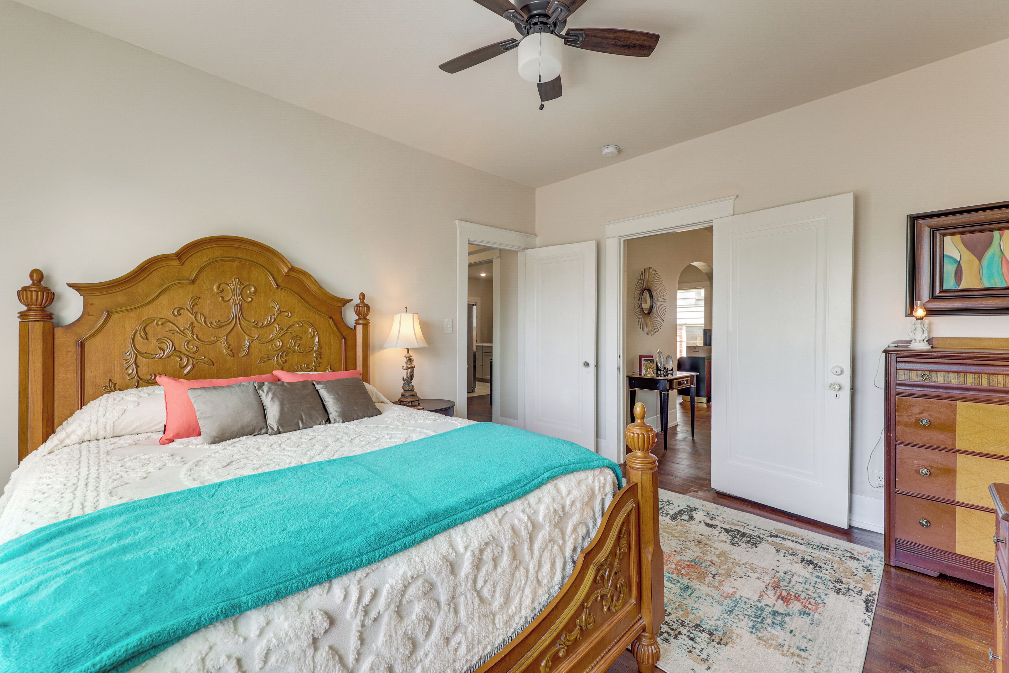 A bedroom with a wooden headboard, a bed with a teal blanket, a nightstand with a lamp, a dresser, and a ceiling fan.