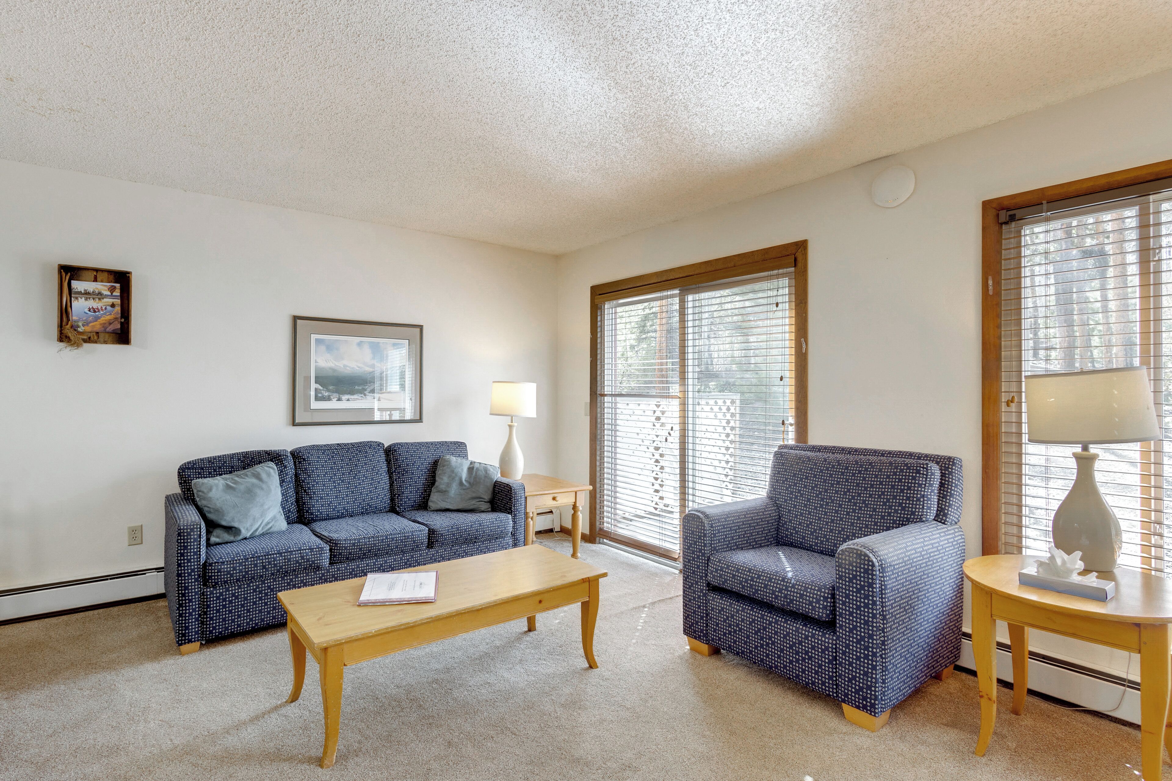 A living room with a blue patterned sofa, a wooden coffee table, a blue armchair, a small wooden side table, and a window with blinds.