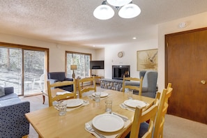 A dining area with a wooden table set for four, surrounded by wooden chairs. There is a sofa and a television in the background. - Walk to Trails & Hideaway Park! Main Street Condo (Winter Park)