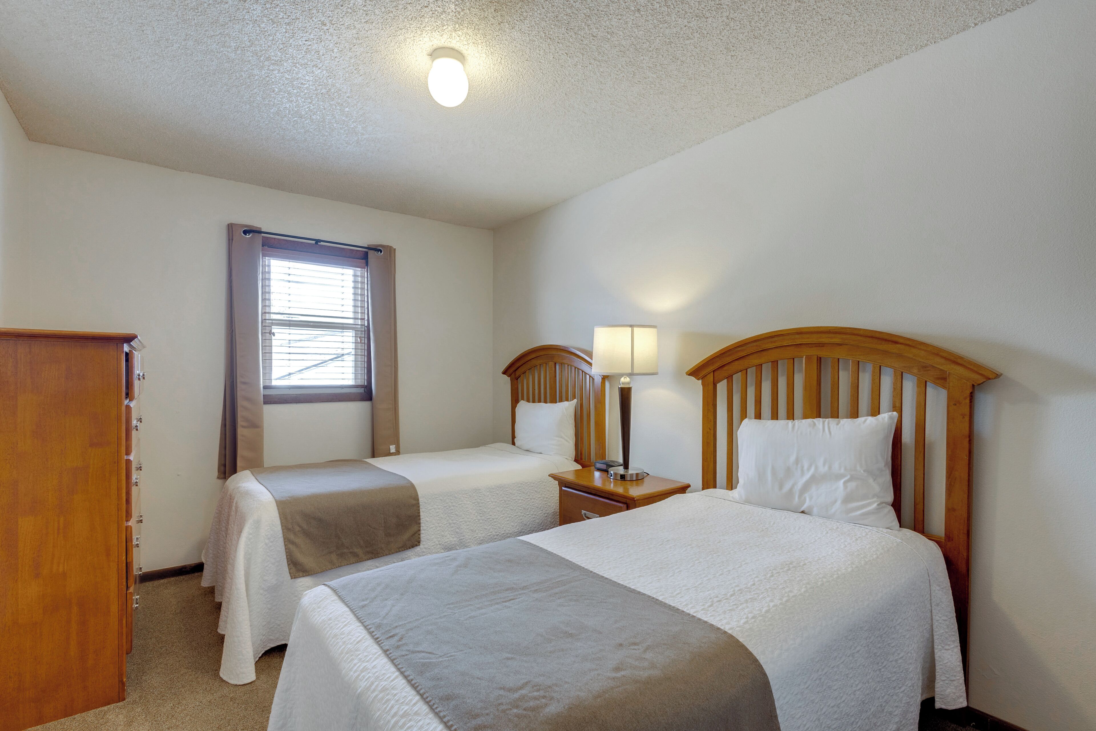 A hotel room with two beds, a wooden dresser, a nightstand with a lamp, and a window with blinds.