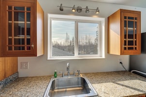 Una cocina con encimeras de granito, pileta de acero inoxidable, gabinetes de madera, un ventanal con vista a árboles y un lámpara en el techo.