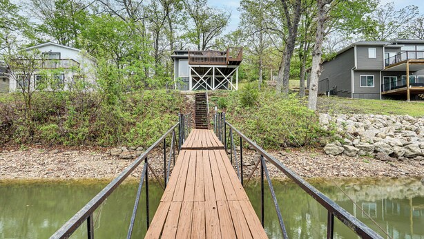 A wooden deck leading to a house with a deck area, surrounded by trees and a body of water. - Dock + Swim Platform! Osage Beach Waterfront Home (Osage Beach)