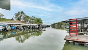 A calm waterway with docked boats, a wooden pier, and a covered area with a red ladder. - Dock + Swim Platform! Osage Beach Waterfront Home (Osage Beach)