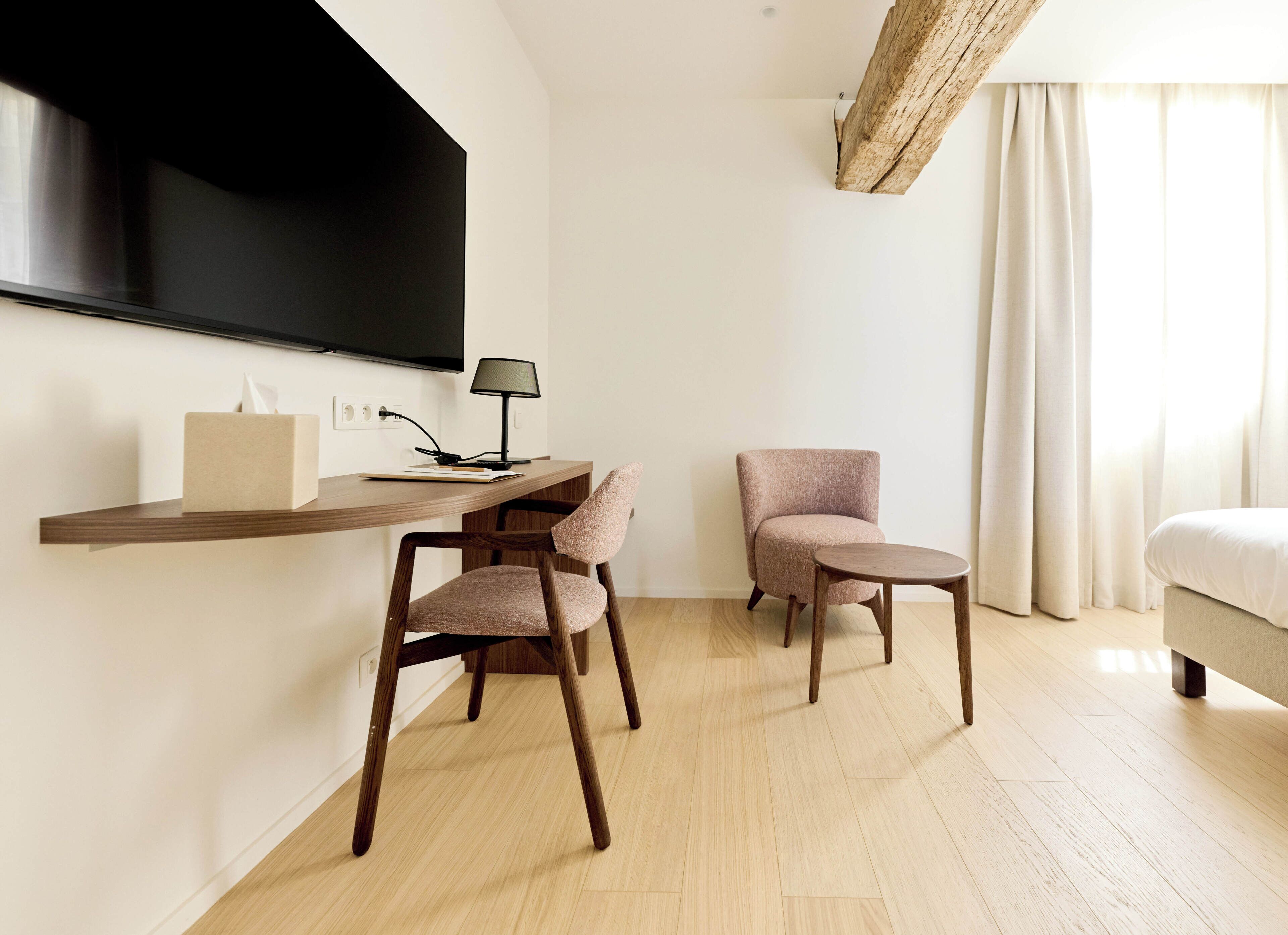 A modern hotel room with a flat-screen TV mounted on the wall, a wooden desk with a lamp, two chairs, and a bed with a white comforter.