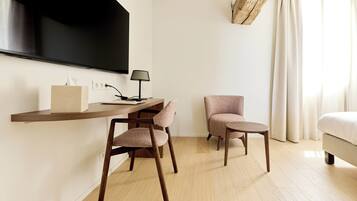 A modern hotel room with a flat-screen TV mounted on the wall, a wooden desk with a lamp, two chairs, and a bed with a white comforter.
