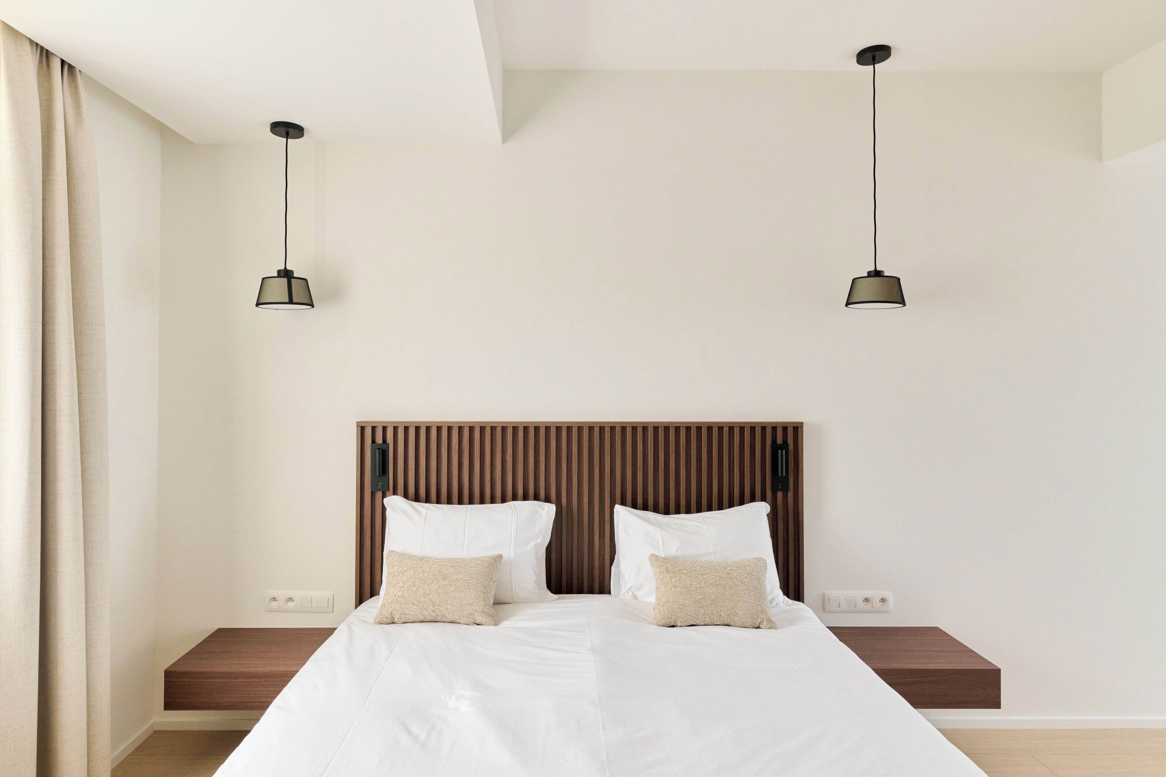 A modern bedroom with a wooden headboard, a large bed with white bedding, two bedside tables, and two pendant lights.