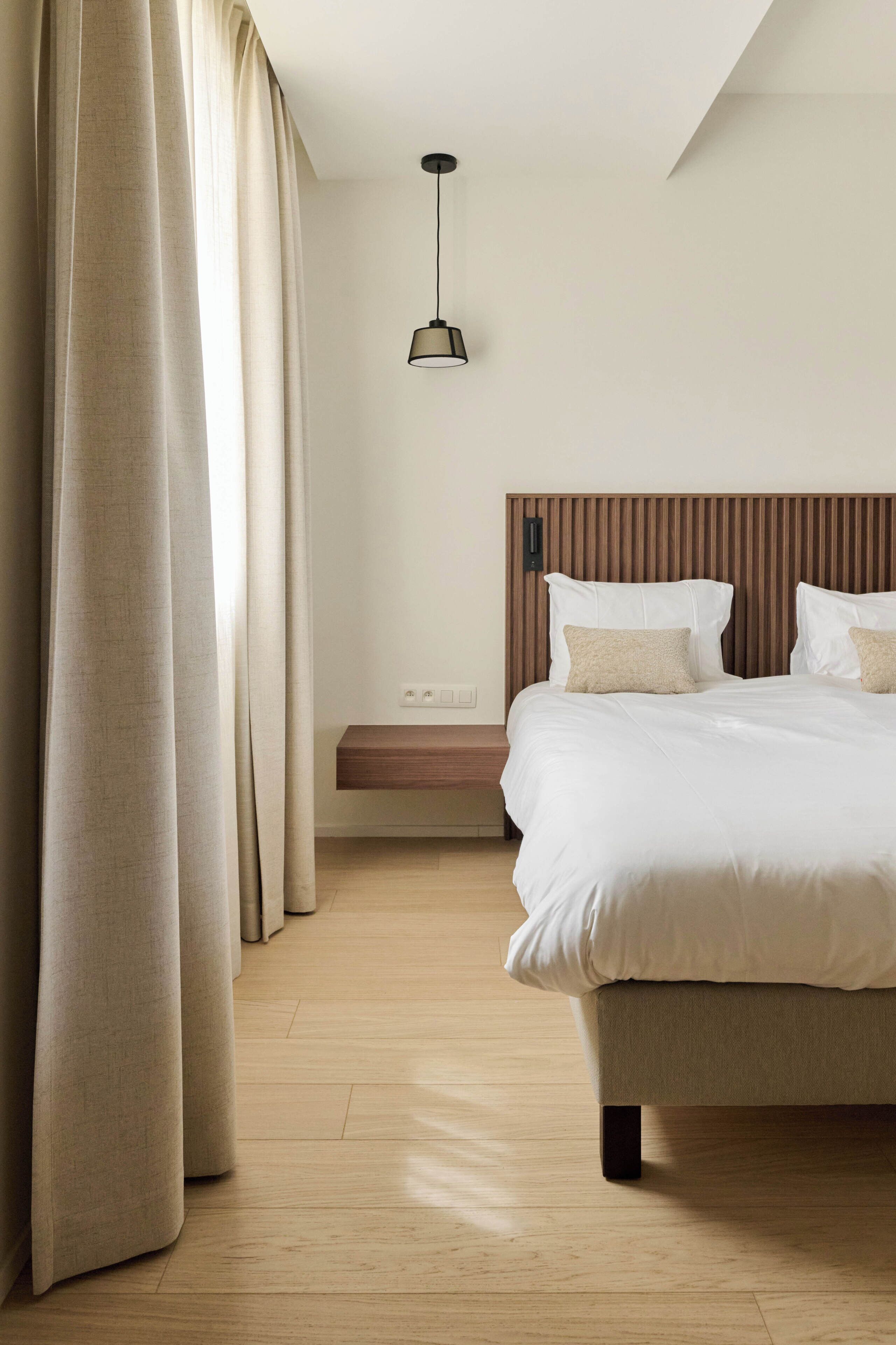 A bedroom with a bed, a bedside table, a hanging light fixture, and a window with curtains.