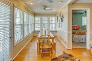 A dining area with a wooden table and chairs, large windows with blinds, a ceiling fan, and a view into a bedroom. - Steps to Shore! Canal-front Escape in Holden Beach (Supply)