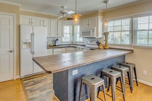 A kitchen with white cabinets, a wooden island, bar stools, a ceiling fan, and a large window. - Steps to Shore! Canal-front Escape in Holden Beach (Supply)