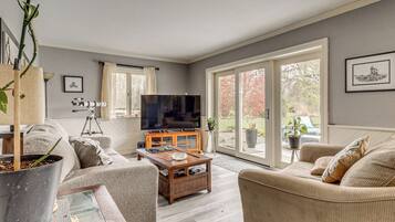 A living room with a sofa, coffee table, and a TV. There is a large window and a door leading outside.