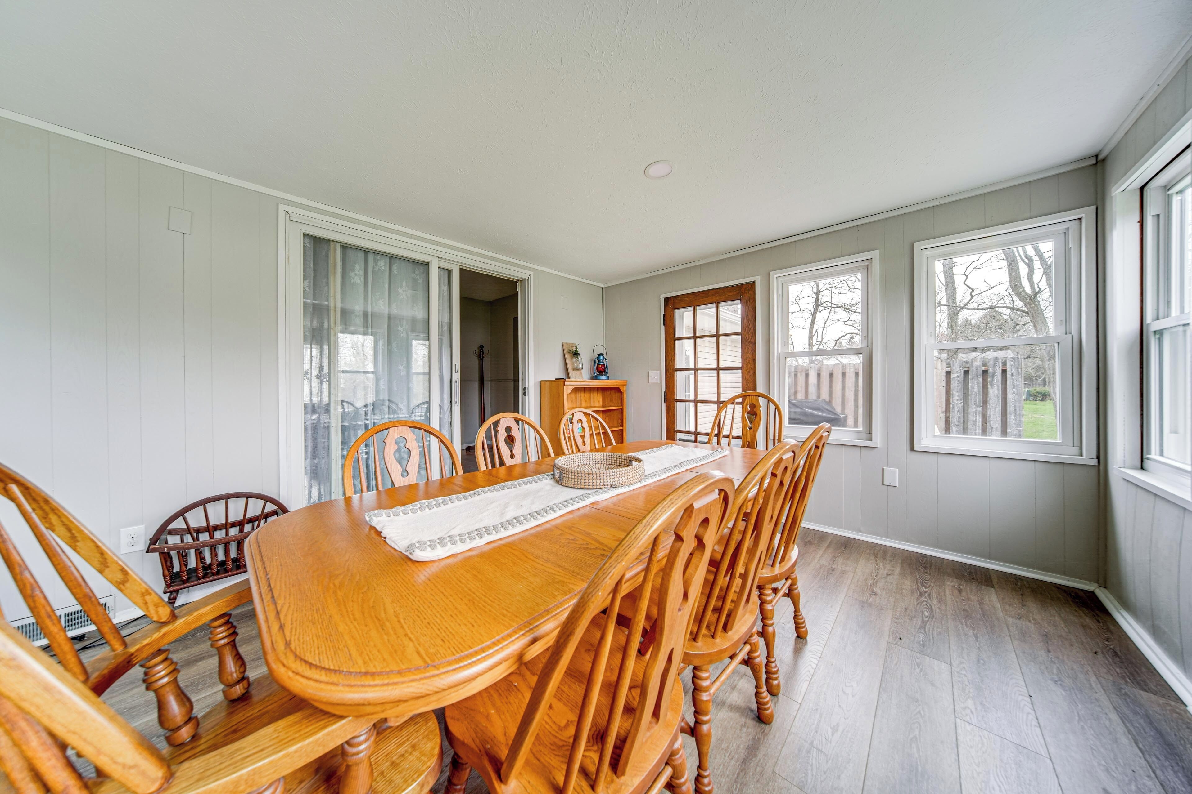A dining room with a wooden table and chairs, large windows, and a view of the outside.