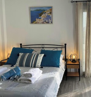 A neatly made bed with blue and white bedding, a painting of a coastal scene, and a nightstand with a lamp. - Aegean Aura Santorini Studio (Perissa)