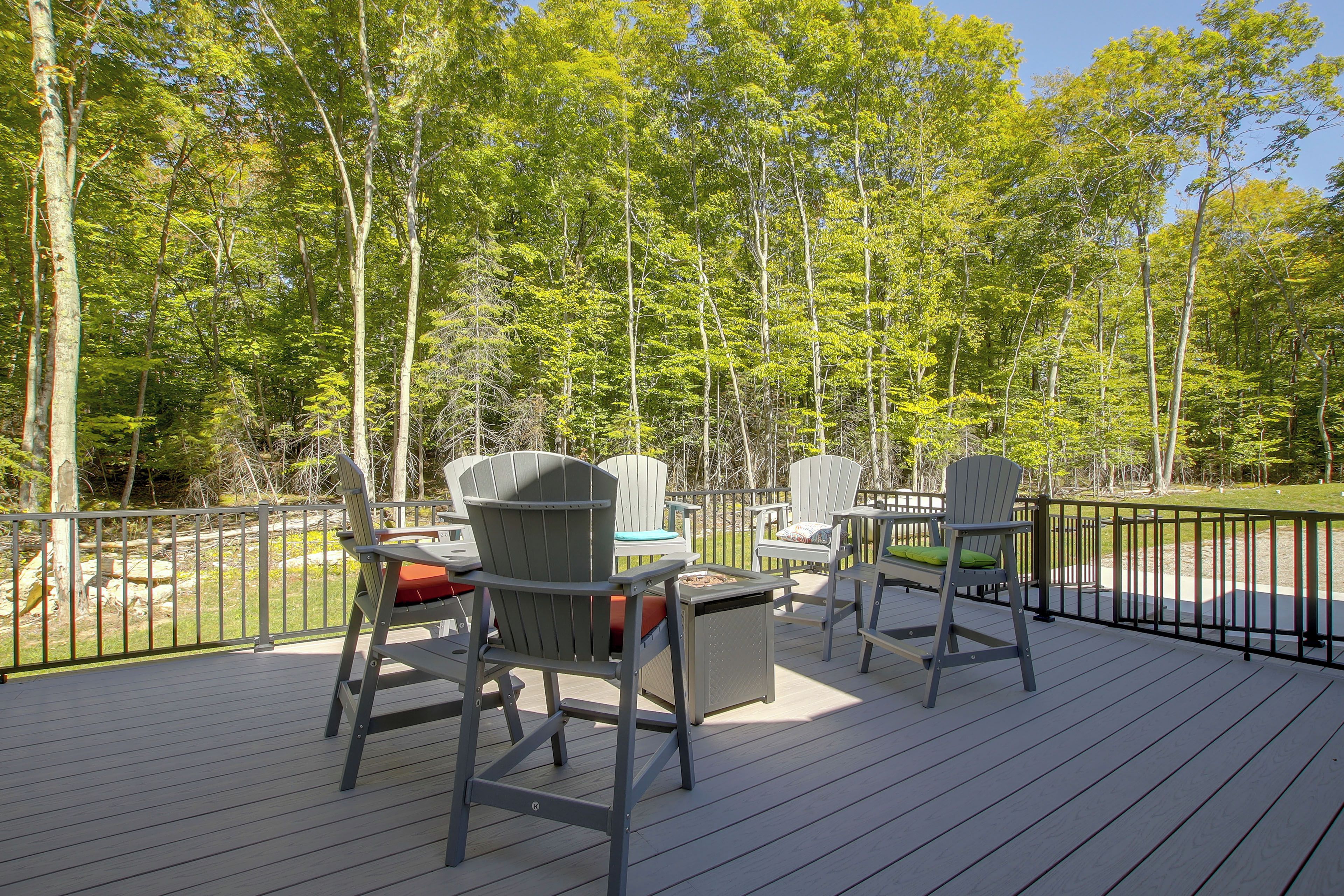 An outdoor deck with Adirondack chairs and a table, surrounded by a metal railing, overlooking a forest.