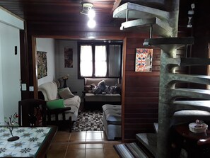 A living room with a sofa, a small table, a decorative plant, and a window with curtains. - Wooden house. (Campos do Jordão)