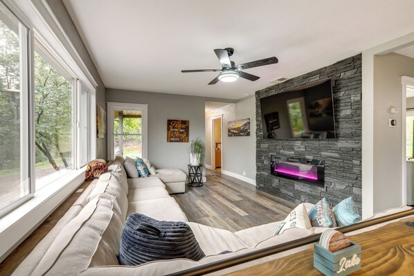 A modern living room with a sectional sofa, a ceiling fan, a stone fireplace, and large windows.