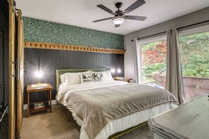 A bedroom with a bed, bedside tables, a ceiling fan, a window with curtains, and a view of greenery outside.