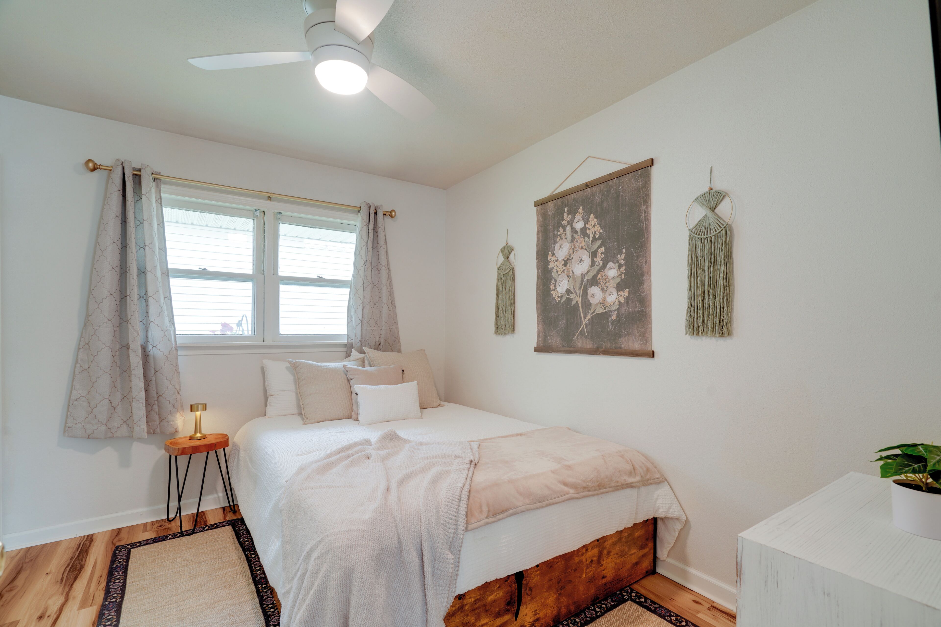 Une chambre à coucher comprenant un lit, une table de chevet avec une lampe, un ventilateur de plafond, une fenêtre avec des rideaux et des éléments de décoration murale.