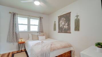 Une chambre à coucher comprenant un lit, une table de chevet avec une lampe, un ventilateur de plafond, une fenêtre avec des rideaux et des éléments de décoration murale.